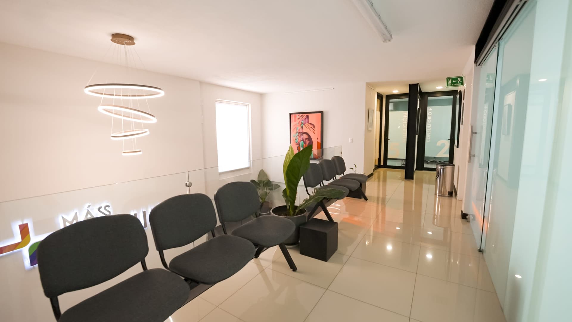 Sala de espera de +Máss Clinik con el logo iluminado y sillas junto a un pasillo de cristal
